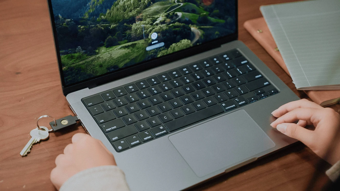 Prijenosnik s prijavnim zaslonom macOS-a i sigurnosnim ključem YubiKey u lijevom priključku, na drvenom stolu uz bilježnicu